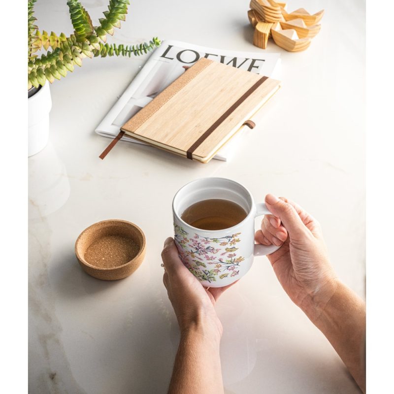 Hands holding floral mug on desk with notebook.